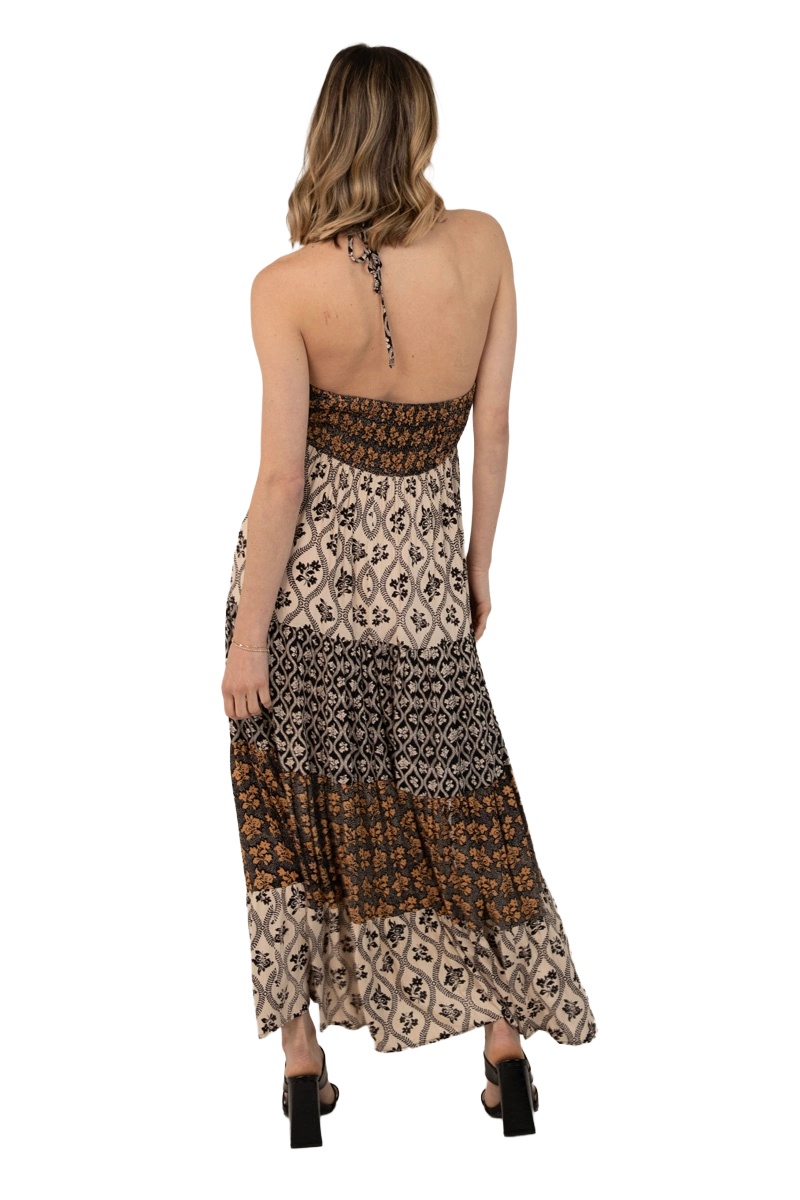 Back view of a mixed‑print halter‑neck maxi dress with an open back, tie‑neck halter strap, and tiered skirt in earthy abstract and geometric prints.